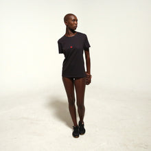 Load image into Gallery viewer, A model stands in a studio wearing a black short-sleeve t-shirt with a small red bunnies logo on the left chest, and black sneakers against a plain off-white background.
