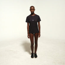 Load image into Gallery viewer, A model stands facing forward in a black short-sleeve t-shirt with a small white bunnies logo on the left chest, paired with black shorts and black sneakers, against a plain off-white studio background.
